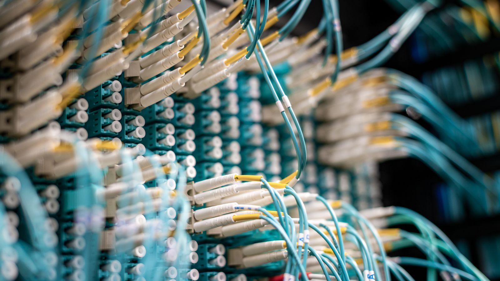 A long-exposure photograph of server room cables glowing pale blue, with a single fibre strand pulled taut across the frame.